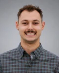 Jonah-paxton | Safe Fleet | Safe Fleet Portrait of a Smiling Man Wearing a Blue-gray Plaid Shirt Against a Neutral Gray Background.