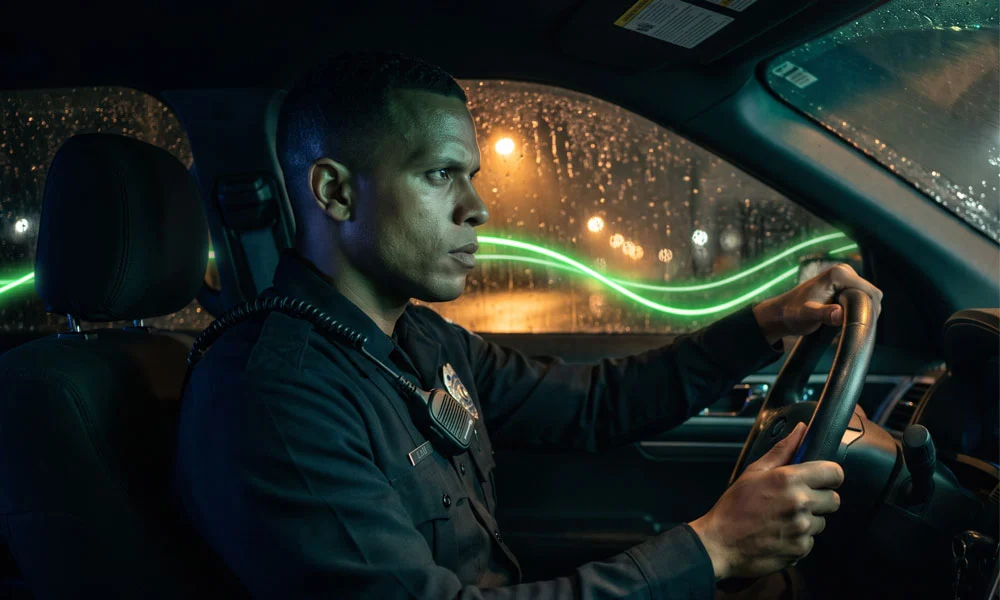 Law-enforcement-blog-3 | Safe Fleet | Safe Fleet Uniformed Police Officer Driving a Patrol Car at Night, Hands on the Steering Wheel, Rain on the Windshield Outside.