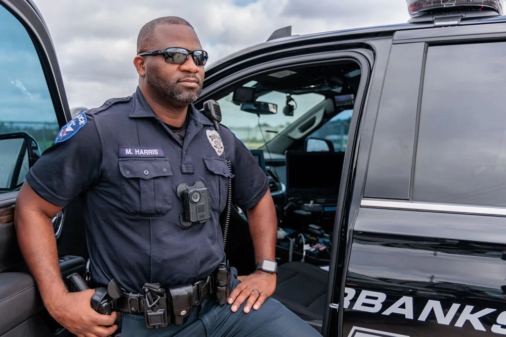 Police-officer-law-enforcement-with-body-camera | Safe Fleet Police Officer in Uniform Leaning Against an Open Police Suv, Wearing Sunglasses and a Duty Belt with Radio and Gear.