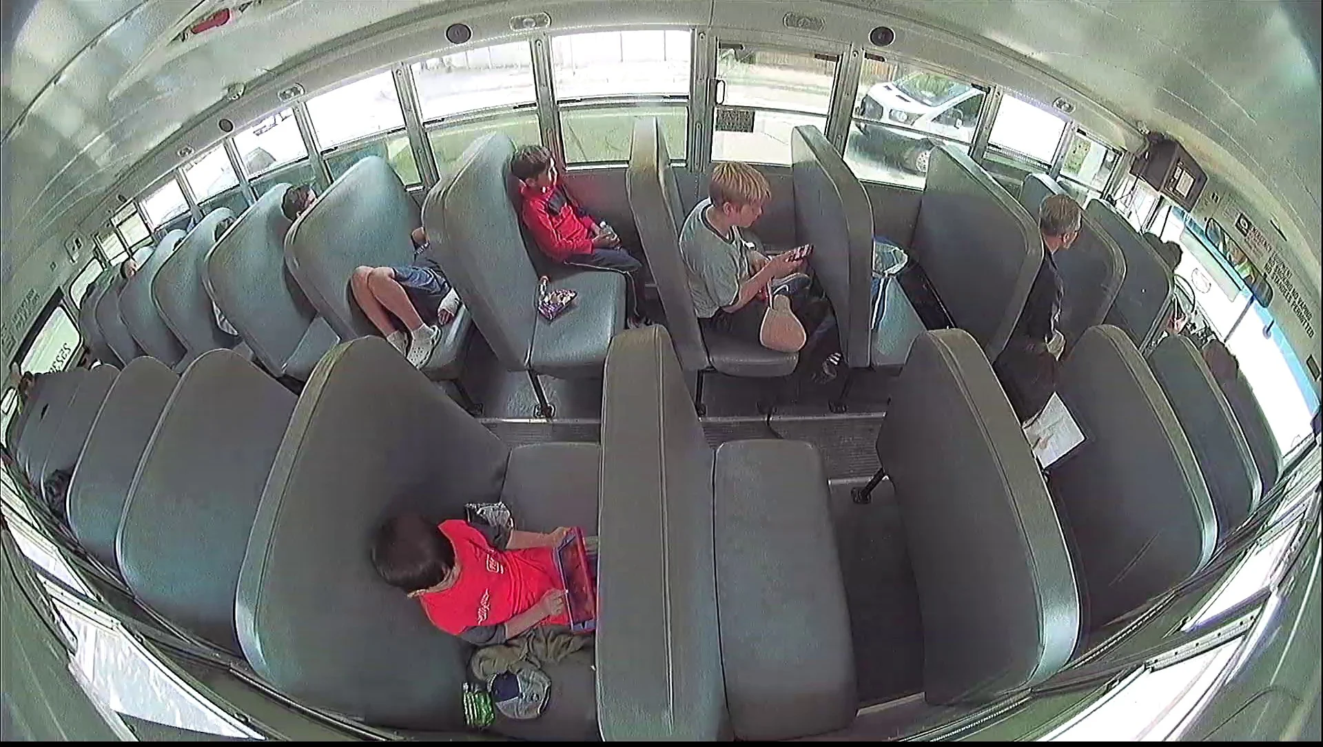 Interior of a School Bus with Several Students Seated in Gray Seats, Some Looking at Phones and Bags on the Floor by the Aisles.