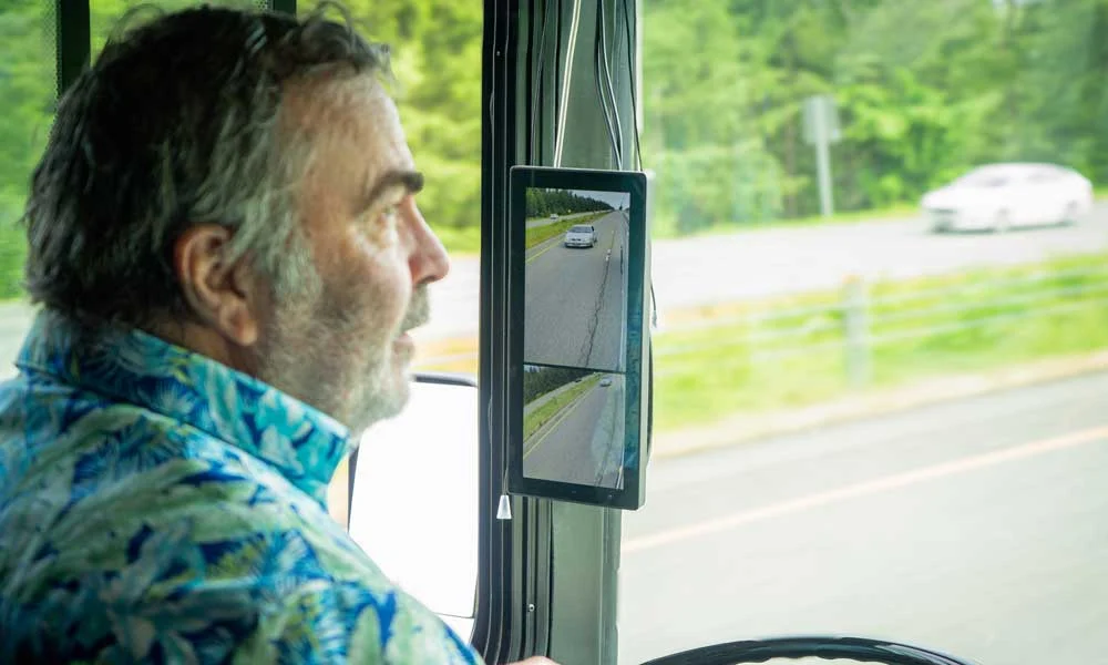 The-ultimate-guide-to-protect-students-in-the-school-bus-danger-zone-blog-article-1 | Safe Fleet | Safe Fleet Man in a Colorful Shirt Driving a Bus, Looking Ahead with a Mounted Dual-screen Camera Display Showing Road Views on the Dashboard.