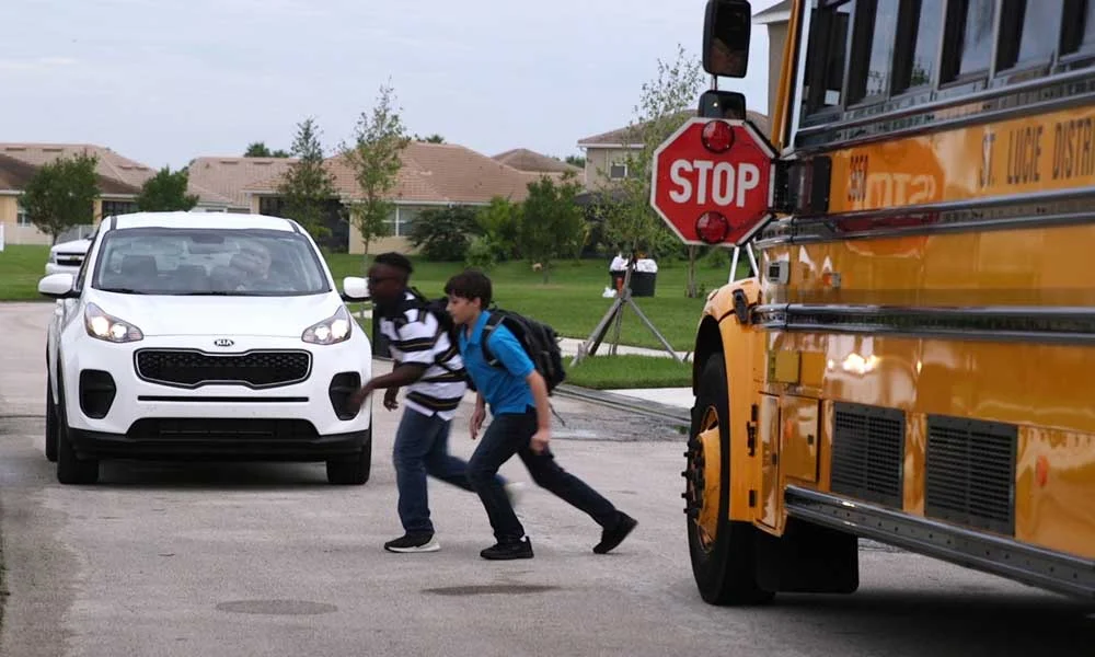Two students cross the street in front of a parked white car with a yellow school bus nearby and a STOP sign in view.