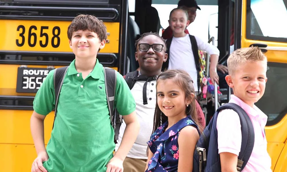 The-ultimate-guide-to-protect-students-in-the-school-bus-danger-zone-blog-article | Safe Fleet | Safe Fleet Group of Smiling Kids with Backpacks Standing in Front of a Yellow School Bus After Getting off the Bus.