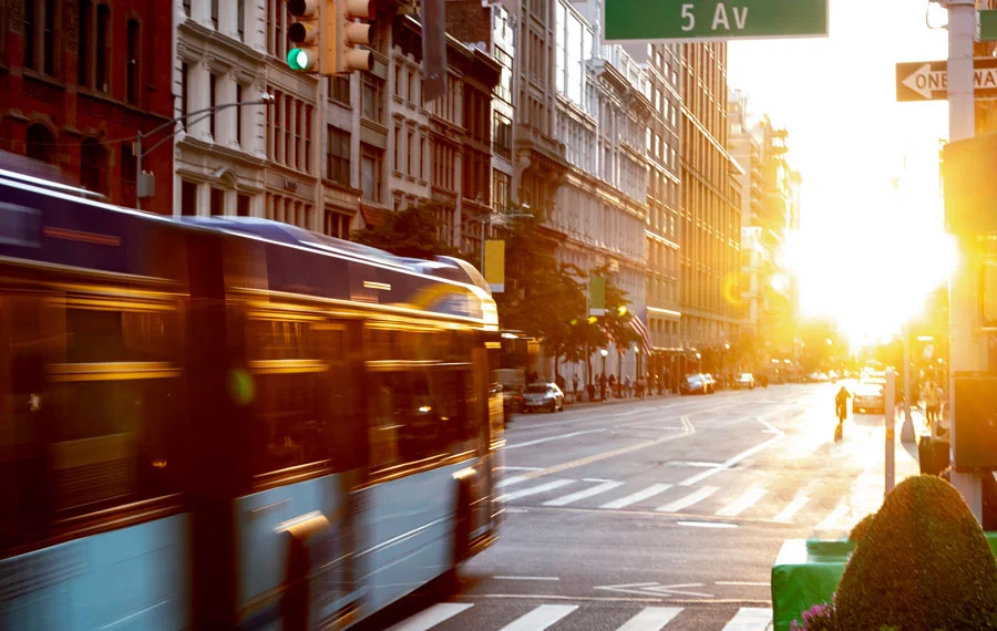Wireless-video-downloading-transit-bus | Safe Fleet | Safe Fleet Blue City Bus in Motion on a Sunlit Downtown Street with Tall Buildings and Traffic in the Distance.